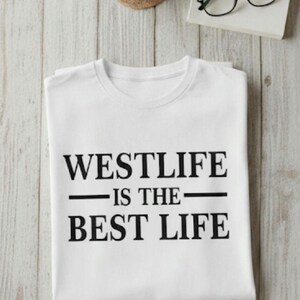 May include: White t-shirt with the text "WESTLIFE IS THE BEST LIFE" in black. A cup of coffee, a coaster, a book, and glasses are on a light wood surface.