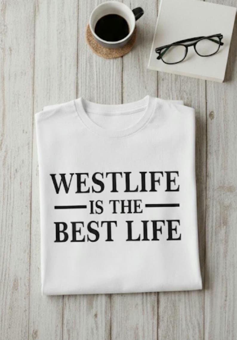 May include: White t-shirt with the text "WESTLIFE IS THE BEST LIFE" in black. A cup of coffee, a coaster, a book, and glasses are on a light wood surface.