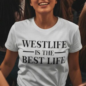 May include: A white t-shirt with the text "WESTLIFE IS THE BEST LIFE" in black. The shirt is knotted at the waist and worn with blue jeans. The person wearing the shirt is smiling.