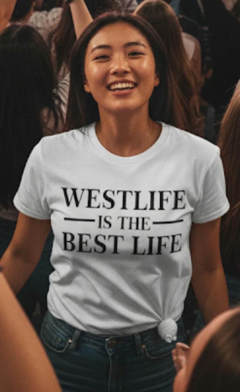 May include: A white t-shirt with the text "WESTLIFE IS THE BEST LIFE" in black. The shirt is knotted at the waist and worn with blue jeans. The person wearing the shirt is smiling.
