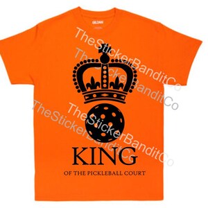 May include: Orange t-shirt with a black crown and a black pickleball. The text "KING OF THE PICKLEBALL COURT" is printed below the pickleball.