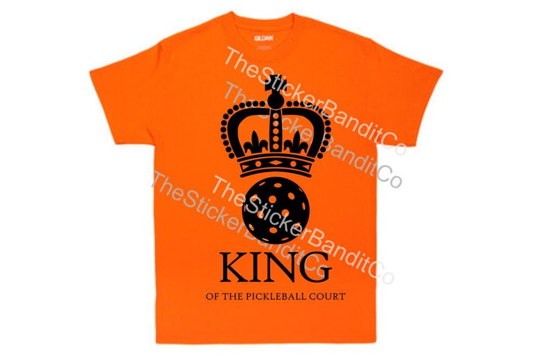 May include: Orange t-shirt with a black crown and a black pickleball. The text "KING OF THE PICKLEBALL COURT" is printed below the pickleball.