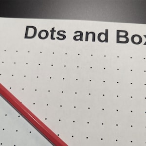 May include: A white sheet of paper with the words "Dots and Boxes" printed at the top. A red pencil is lying on the paper, which is covered in a grid of small black dots.