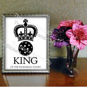 May include: A black and white image of a pickleball with a crown on top. The text "KING OF THE PICKLEBALL COURT" is below the pickleball.