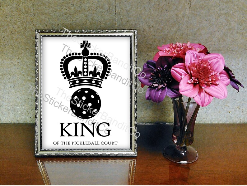 May include: A black and white image of a pickleball with a crown on top. The text "KING OF THE PICKLEBALL COURT" is below the pickleball.