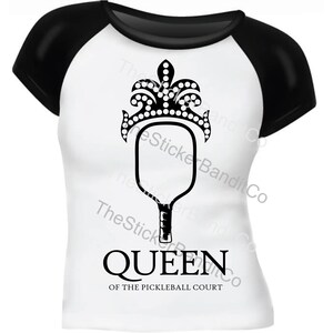 May include: A white t-shirt with black sleeves featuring a black and white illustration of a pickleball paddle with a crown on top. The text "QUEEN OF THE PICKLEBALL COURT" is printed below the paddle.