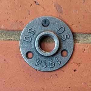 May include: A collection of black metal pipe flanges with a circular design and multiple screw holes. One flange is mounted on a red brick wall. The flange has the text "DS OS 1914" stamped on it.