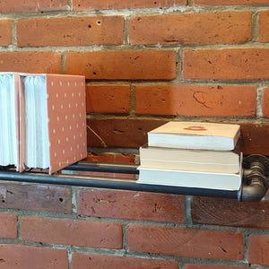 May include: A black metal pipe shelf mounted on a red brick wall, holding books. Two pink polka-dotted books stand upright, with a stack of books to the right. The shelf is a decorative storage solution.