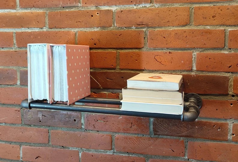 May include: A black metal pipe shelf mounted on a red brick wall, holding books. Two pink polka-dotted books stand upright, with a stack of books to the right. The shelf is a decorative storage solution.