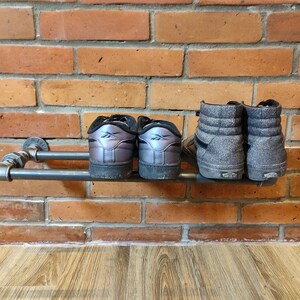 May include: A metal pipe shoe rack mounted on a brick wall. Two pairs of shoes are displayed: low-top sneakers with iridescent uppers and high-top sneakers in a speckled gray fabric.