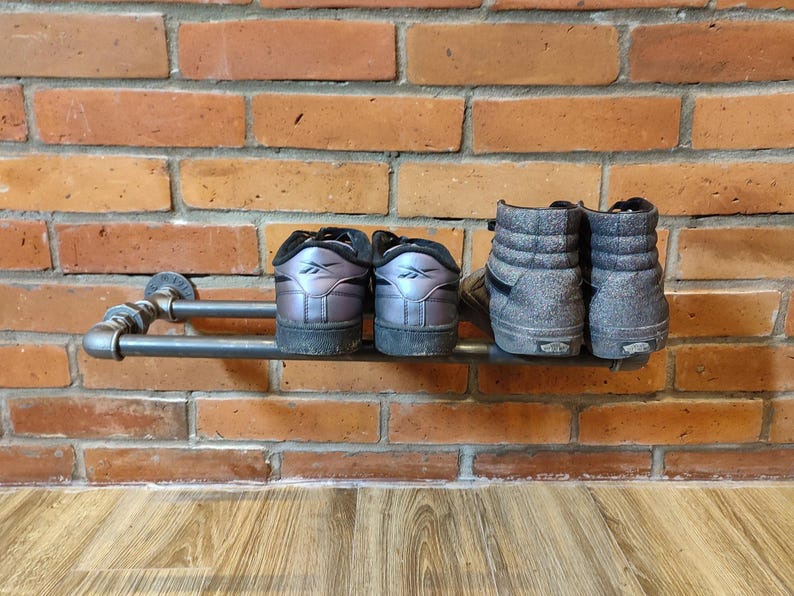 May include: A metal pipe shoe rack mounted on a brick wall. Two pairs of shoes are displayed: low-top sneakers with iridescent uppers and high-top sneakers in a speckled gray fabric.