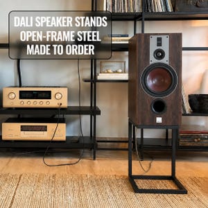 Può includere: Un altoparlante marrone su un supporto in acciaio nero a telaio aperto. L'altoparlante ha un pannello argentato con un altoparlante nero e un centro rosso. Il testo "DALI SPEAKER STANDS OPEN-FRAME STEEL MADE TO ORDER" è in alto a sinistra.