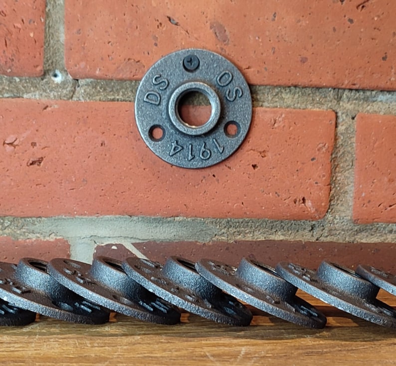 May include: A collection of black metal pipe flanges with a circular design and multiple screw holes. One flange is mounted on a red brick wall. The flange has the text "DS OS 1914" stamped on it.