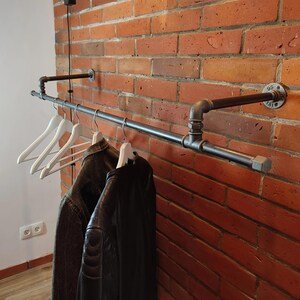 May include: A black metal pipe clothing rack mounted on a red brick wall. White wooden hangers hold a dark jacket and a black leather jacket. The industrial design features pipe fittings and a minimalist aesthetic.