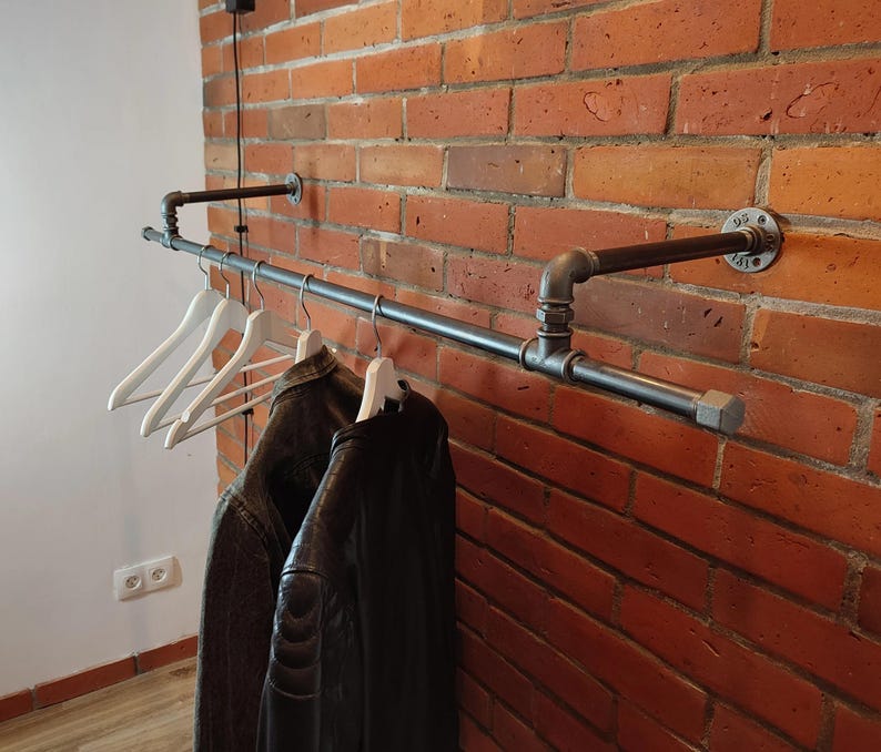 May include: A black metal pipe clothing rack mounted on a red brick wall. White wooden hangers hold a dark jacket and a black leather jacket. The industrial design features pipe fittings and a minimalist aesthetic.