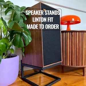 Puede incluir: Un soporte de altavoz de madera con un panel de altavoz negro y una base de metal negro. El texto en el soporte dice "SPEAKER STANDS LINTON FIT MADE TO ORDER". El soporte está junto a una planta en maceta y un mueble de estilo moderno de mediados de siglo.