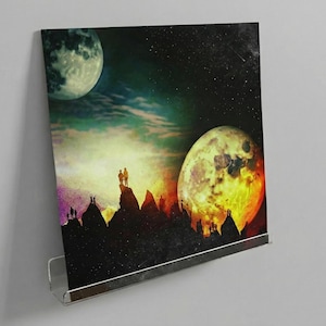 Puede incluir: Impresión enmarcada que representa un paisaje surrealista con dos lunas en un cielo estrellado. Figuras silueteadas se alzan sobre picos montañosos. Las lunas son de un verde pálido y un naranja fuego, creando un contraste dramático.