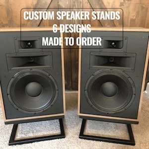 May include: Two black speaker cabinets with light wood frames and black metal stands. The speakers have large circular woofers and text that reads "CUSTOM SPEAKER STANDS 6 DESIGNS MADE TO ORDER". The speakers are on a light-colored carpet.