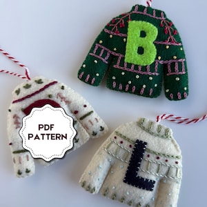 Può includere: Tre decorazioni a forma di maglione in feltro con ganci in spago rosso e bianco. Un maglione verde ha una "B" verde lime, un altro maglione bianco sporco ha una "L" nera e il terzo ha un'etichetta "PDF PATTERN".