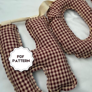 Pode incluir: Uma faixa de tecido com a palavra "HO" em estampa xadrez vichy bordô e creme. As letras são acolchoadas e costuradas com linha creme. A faixa é presa a uma tira de tecido creme. Um adesivo branco com o texto "PDF PATTERN" é visível.