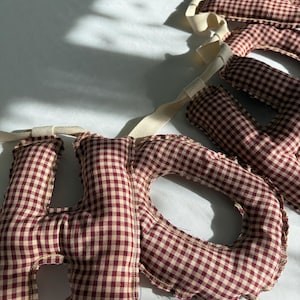 May include: A fabric banner spelling out the word "HOME" in burgundy and cream gingham letters. The letters are sewn with a scalloped edge and attached to a cream-colored ribbon. The banner is displayed on a white surface.