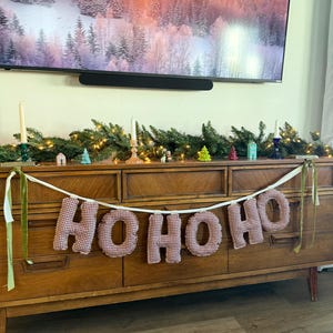 May include: A wooden dresser with a "HO HO HO" banner made of red and white checkered fabric. The dresser is decorated with a garland of greenery, candles, and miniature houses and trees, creating a festive holiday display.