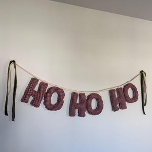 May include: A festive banner with the words "HO HO HO" in burgundy and white checkered fabric letters. The letters are strung on a thin, off-white cord, with green and cream-colored ribbons at each end.