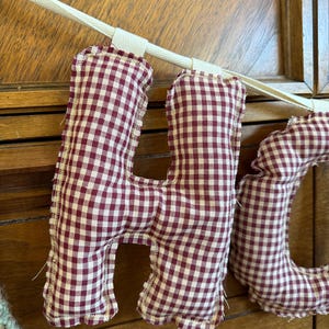 May include: A close-up of a decorative banner spelling out the word "HO" in burgundy and white gingham fabric letters. The letters are sewn with a cream-colored thread and attached to a cream-colored ribbon.