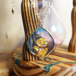 Peut inclure: Un collier en chaîne dorée avec un pendentif représentant une image en résine peinte à la main d'un robot aux yeux bleus. Le pendentif est sur un fond bleu et rose.