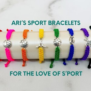 May include: A set of 10 colorful string bracelets with different sports-themed charms. The charms include a basketball, soccer ball, volleyball, tennis racket, hockey sticks, and a football. The bracelets are arranged on a white surface with the text "ARI'S SPORT BRACELETS" and "FOR THE LOVE OF S'PORT" above and below the bracelets. A teal ribbon is in the upper right corner of the image.