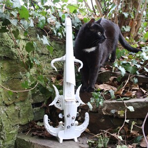 K&ouml;nnte beinhalten: Eine wei&szlig;e elektrische Geige mit filigranen Details und eine schwarze Katze, die auf einer Steinstufe mit gr&uuml;nem Efeu sitzt.