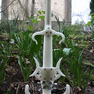 May include: A white 3D printed electric violin with intricate details and a unique design. The violin is standing on a stand and is surrounded by green grass and brown leaves.