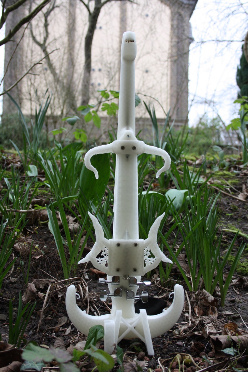 May include: A white 3D printed electric violin with intricate details and a unique design. The violin is standing on a stand and is surrounded by green grass and brown leaves.