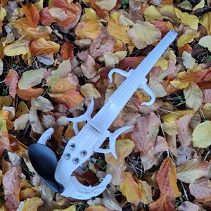 Puede incluir: Un viol&iacute;n el&eacute;ctrico blanco con un dise&ntilde;o &uacute;nico, que descansa sobre un lecho de hojas de oto&ntilde;o ca&iacute;das. El viol&iacute;n tiene una barbilla negra y un cuerpo blanco con detalles intrincados.
