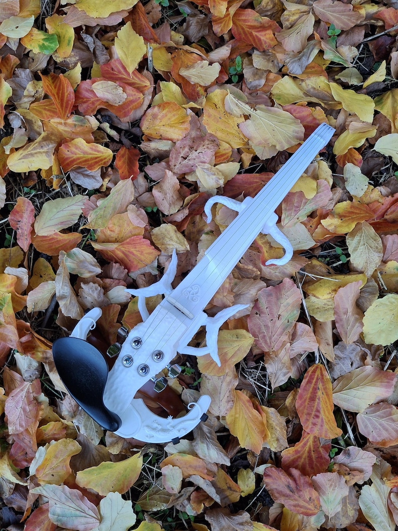 Puede incluir: Un viol&iacute;n el&eacute;ctrico blanco con un dise&ntilde;o &uacute;nico, que descansa sobre un lecho de hojas de oto&ntilde;o ca&iacute;das. El viol&iacute;n tiene una barbilla negra y un cuerpo blanco con detalles intrincados.