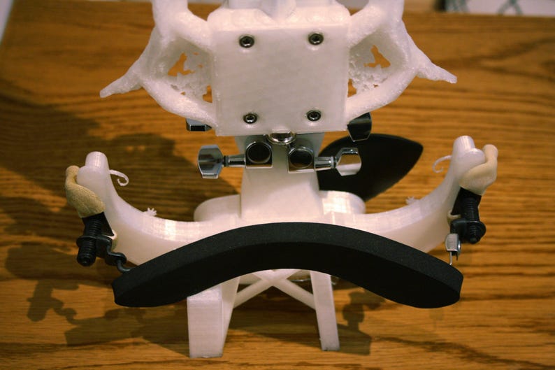 May include: White 3D printed violin shoulder rest with a black foam pad. The shoulder rest has two adjustable arms with metal screws.
