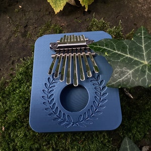Puede incluir: Un kalimba de madera azul con una placa de sonido de metal plateado y un diseño decorativo de corona. El kalimba tiene 8 teclas numeradas del 1 al 8.