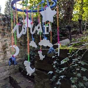 Puede incluir: Un móvil para bebé blanco con un aro azul, cuerdas de colores y varias formas impresas en 3D, incluyendo estrellas, nubes, dinosaurios y una luna creciente. El móvil también incluye la palabra "Lou" en letras blancas.