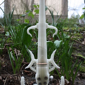 May include: A white 3D printed electric violin with intricate details and a black pick-up. The violin is standing on a black stand in a grassy area.