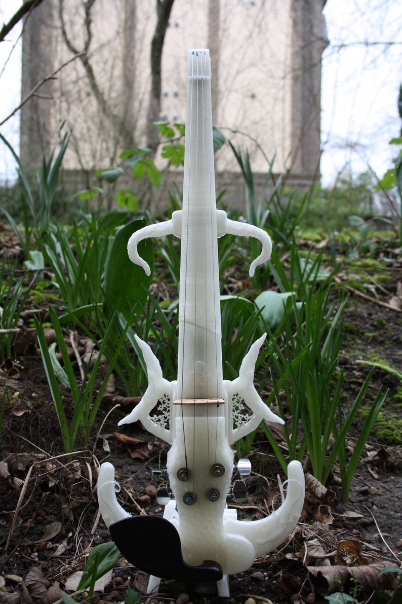 May include: A white 3D printed electric violin with intricate details and a black pick-up. The violin is standing on a black stand in a grassy area.