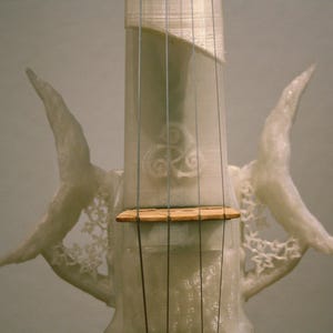 May include: A white 3D printed violin with intricate details and a wooden bridge. The violin has a unique design with two wing-like structures on either side of the neck.