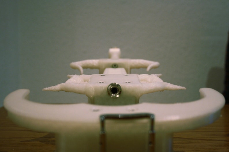 May include: White plastic object with a round hole in the center and two curved arms on either side. The object is designed to hold something in place.