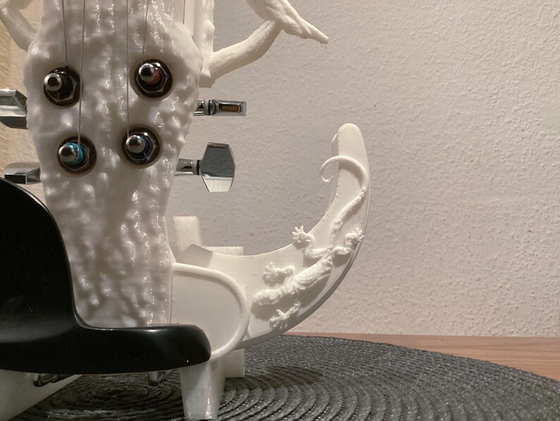 Puede incluir: Guitarra el&eacute;ctrica blanca con un dise&ntilde;o &uacute;nico que presenta un cuerpo de tronco de &aacute;rbol y una cola de lagarto en la parte inferior del cuerpo. La guitarra tiene cuatro clavijas de afinaci&oacute;n y una golpeadora negra.