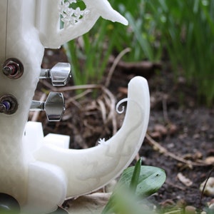 May include: White 3D printed violin with intricate details, including a floral design on the body and a scroll on the neck. The violin has four tuning pegs and is resting on a bed of green grass.