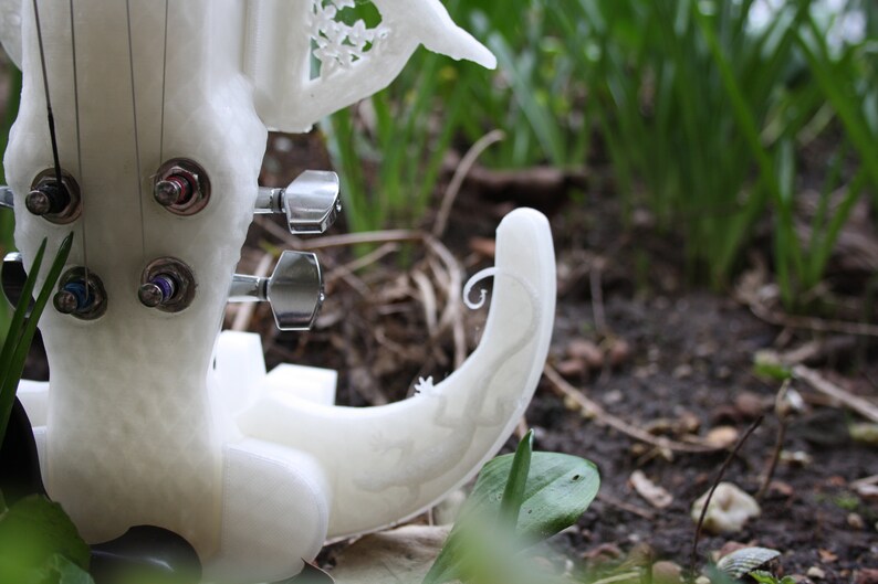 May include: White 3D printed violin with intricate details, including a floral design on the body and a scroll on the neck. The violin has four tuning pegs and is resting on a bed of green grass.