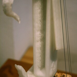 May include: White marble sculpture of a violin with a wooden bridge and strings. The sculpture is on a wooden surface.