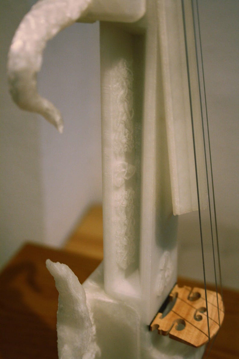 May include: White marble sculpture of a violin with a wooden bridge and strings. The sculpture is on a wooden surface.
