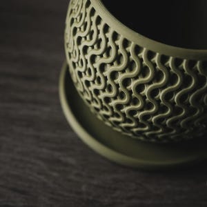 Puede incluir: Una maceta de color verde salvia con forma redondeada y un plato a juego. La superficie exterior presenta un patr&oacute;n de ondas en relieve que crea un aspecto texturizado similar a una rejilla. El logotipo h3LIO es visible abajo a la derecha.