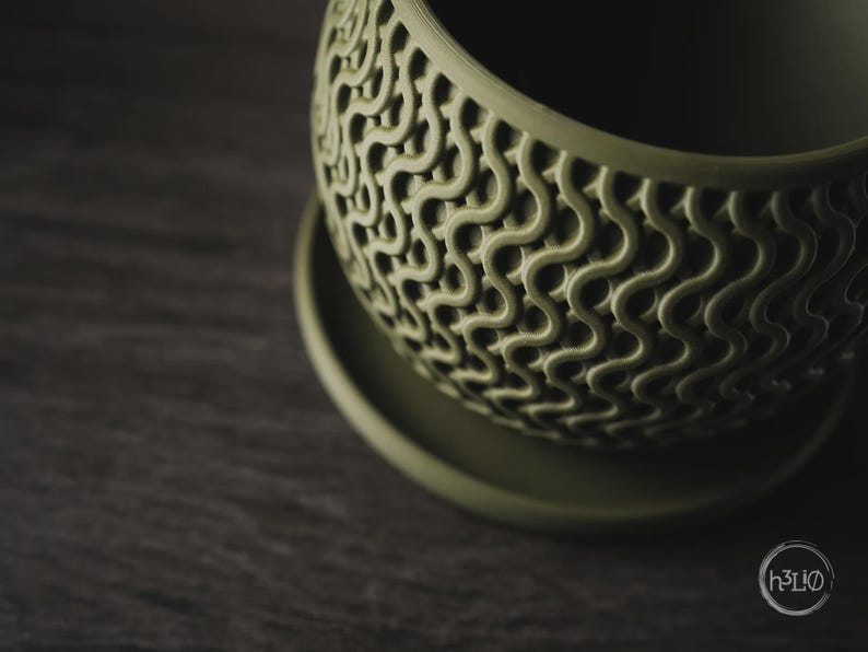 Puede incluir: Una maceta de color verde salvia con forma redondeada y un plato a juego. La superficie exterior presenta un patr&oacute;n de ondas en relieve que crea un aspecto texturizado similar a una rejilla. El logotipo h3LIO es visible abajo a la derecha.