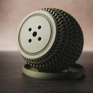 Puede incluir: Un objeto esf&eacute;rico de color verde oliva con una textura compleja de celos&iacute;a ondulada que cubre su superficie. La parte frontal presenta un disco circular plano con cinco peque&ntilde;os orificios circulares espaciados uniformemente.